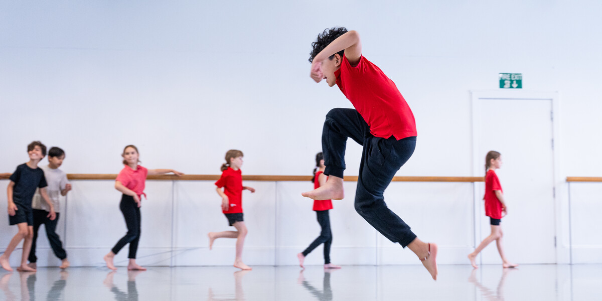 A boy in a red school uniform jumping really high in a dance studio. Other students are lined up in the background.