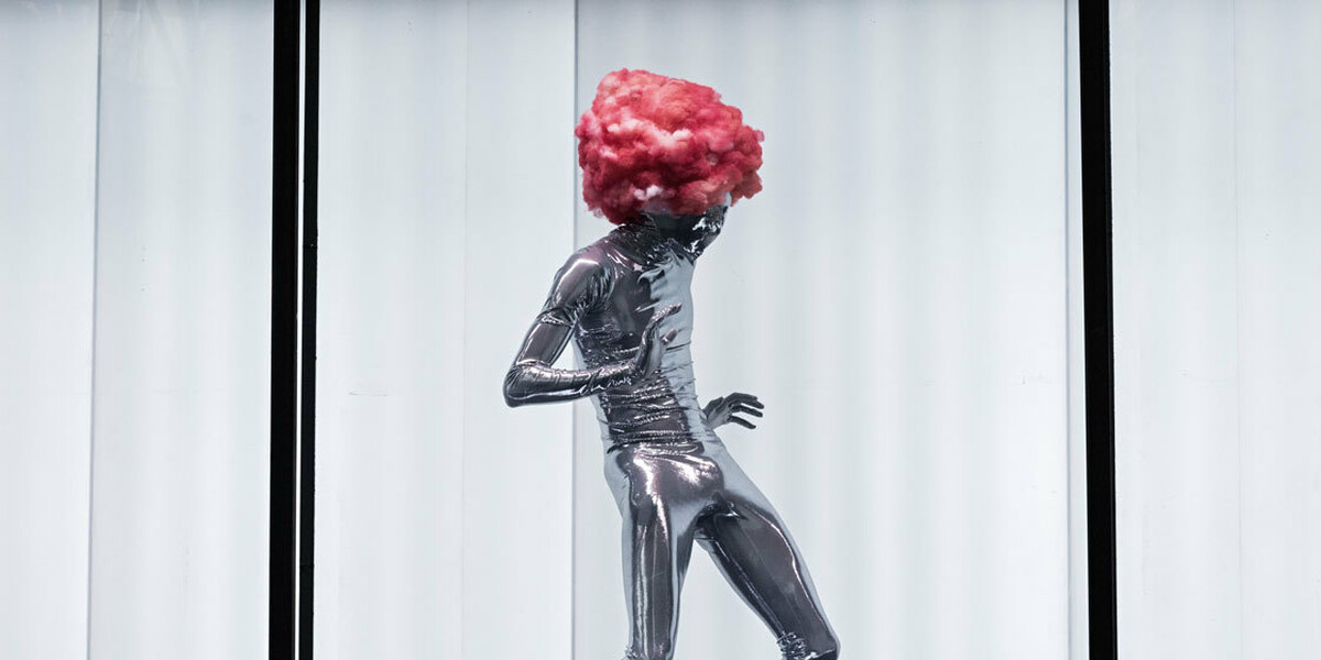 A shiny, silver being wearing a pink candy floss wig dances against the white, lit interior of the shipping container.