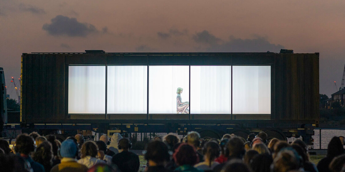 Set against the sky at dusk, a captive audience all wearing headphones are seated in front of a large, dark shipping container, mounted on the bed of a 40ft truck. The long side of the container has been removed revealing a single silver being, crouched on the floor.