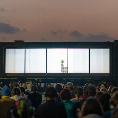 Set against the sky at dusk, a captive audience all wearing headphones are seated in front of a large, dark shipping container, mounted on the bed of a 40ft truck. The long side of the container has been removed revealing a single silver being, crouched on the floor.