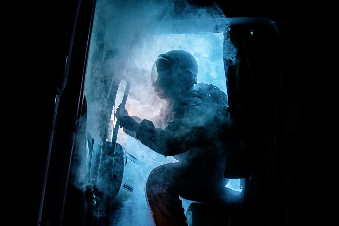 The cabin door of a truck opens, spewing eerie blue smoke. Silhouetted by a bright blue light that cuts through the darkness of night, a figure wearing an astronaut uniform begins to emerge.