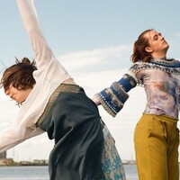 Image of two dancers dancing by the river. In the background are some high rise buildings and a view of the river. One dancer is leaning over to the left with their hands in the air above their heads. They are wearing a white long-sleeved crop top and a bluish dress. The other dancer is standing beside and wears yellow trousers and a patterned top.