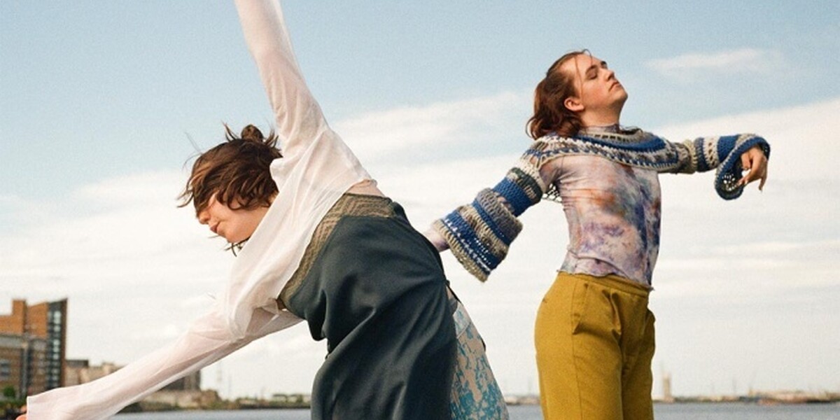 Image of two dancers dancing by the river. In the background are some high rise buildings and a view of the river. One dancer is leaning over to the left with their hands in the air above their heads. They are wearing a white long-sleeved crop top and a bluish dress. The other dancer is standing beside and wears yellow trousers and a patterned top.