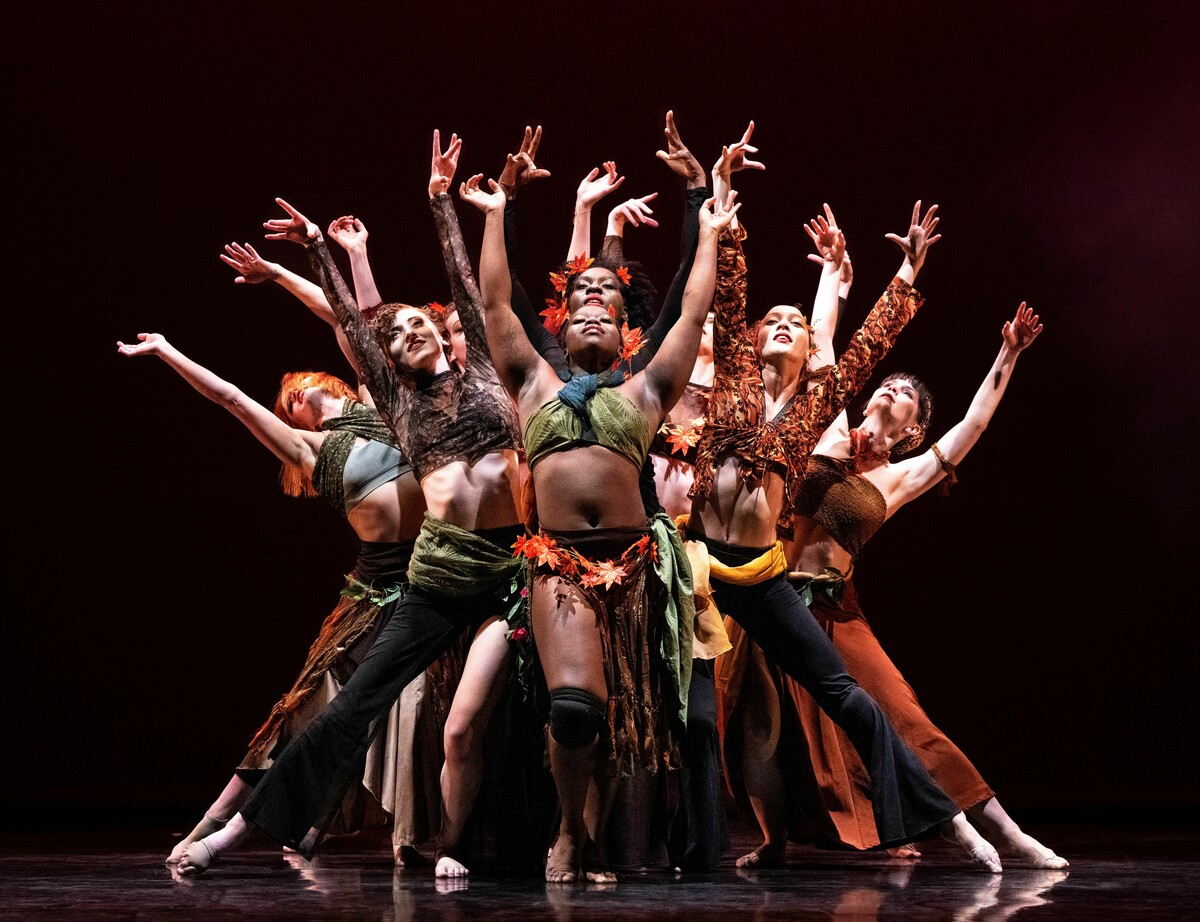 A group of dancers performing onstage. They wear autumnal colours and leaf garlands, and their arms are stretched out above them.