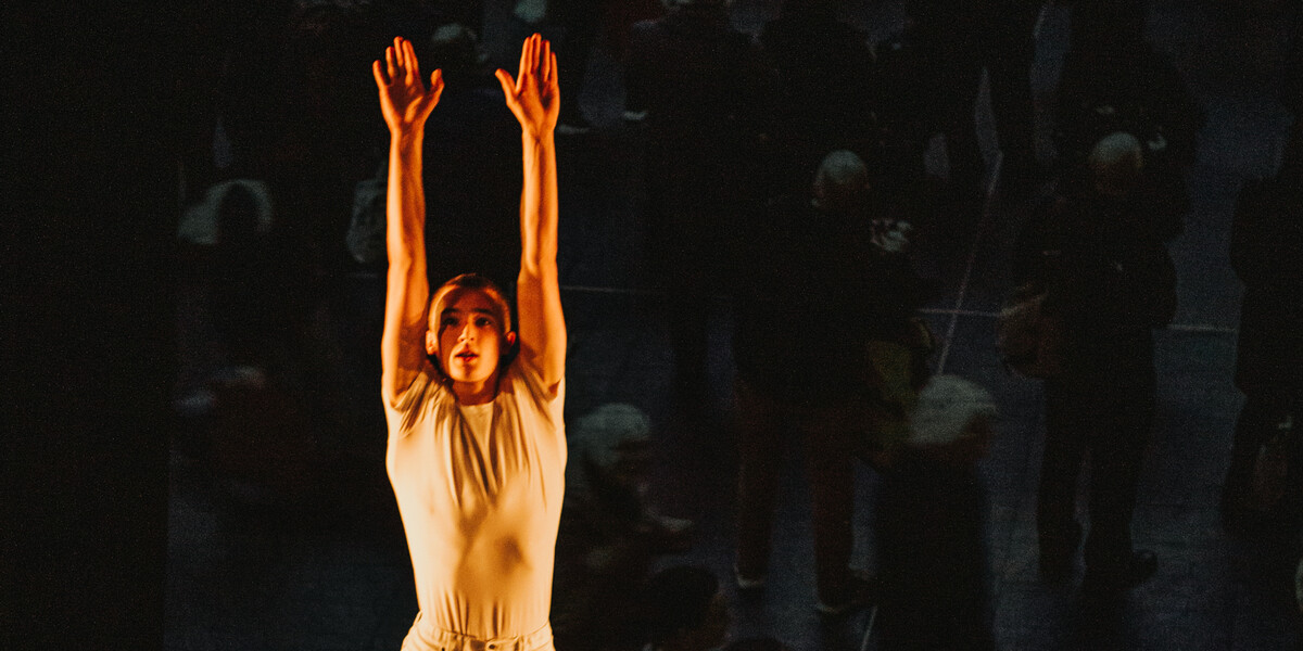 Two dancers, one rolling across the floor, the other jumping over them with their arm stretched to the ceiling and knees bent. They are both wearing white.