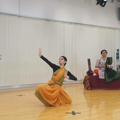 2 Bharatanatyam dancers in a dance studio wearing saris, one is kneeling on the floor, one is instructing. There is a musician in the background.