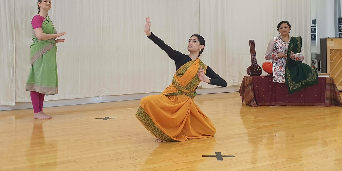 2 Bharatanatyam dancers in a dance studio wearing saris, one is kneeling on the floor, one is instructing. There is a musician in the background.
