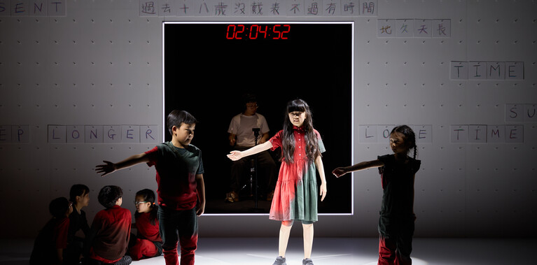 A photo of a cohort of child performers dancing on stage with an adult drummer sat in the background with a motion sensor drumkit and headphones