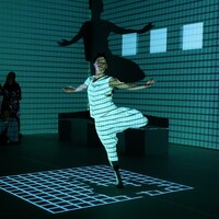 A dancer in a white vest and trousers pirouettes in a dark room, a grid of light projected onto him and the walls behind him. Audience members can be seen around the frame of the image.