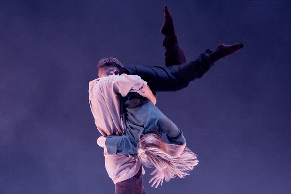 A dancer wearing a long sleeved beige top is lifting another dancer upside down, holding onto them by their waist. The other dancer is wearing black trousers and a dark grey t-shirt. They twist their body and hold the other person's waist with their right arm, whilst bending their left arm above their head and pointing their legs diagonally upwards.