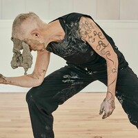 A woman with short hair is wearing a black tank top and trousers has a large chunk of clay on her face and is leaning over with her elbow on her knee.