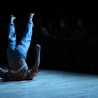 Image of a dancer on their shoulders with their legs up in the air. The light makes the image more blue. The dancer is wearing a shirt and jeans.
