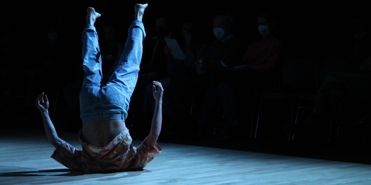 Image of a dancer on their shoulders with their legs up in the air. The light makes the image more blue. The dancer is wearing a shirt and jeans.