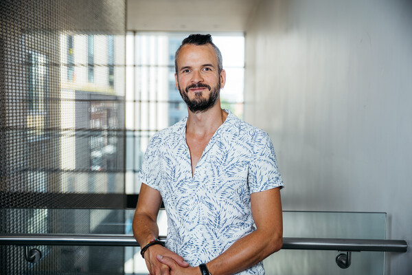 A man with short black hair and a black beard looks at the camera with a slight smile. He is wearing a light blue and white patterned shirt and is leaning on a railing with his hand clasped together