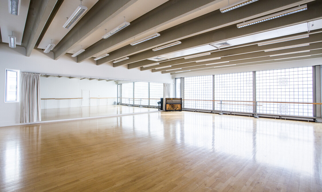 Image of an empty studio with widows on the right hand side and a ballet barre across the windows and mirrors on the left wall. There is a piano in the corner