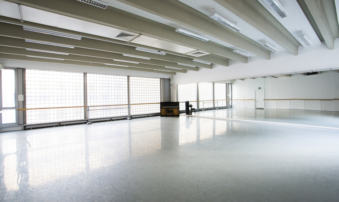 Image of an empty studio with mirrors on the right hand side and widows on the left with ballet barres across the window and a piano in the corner