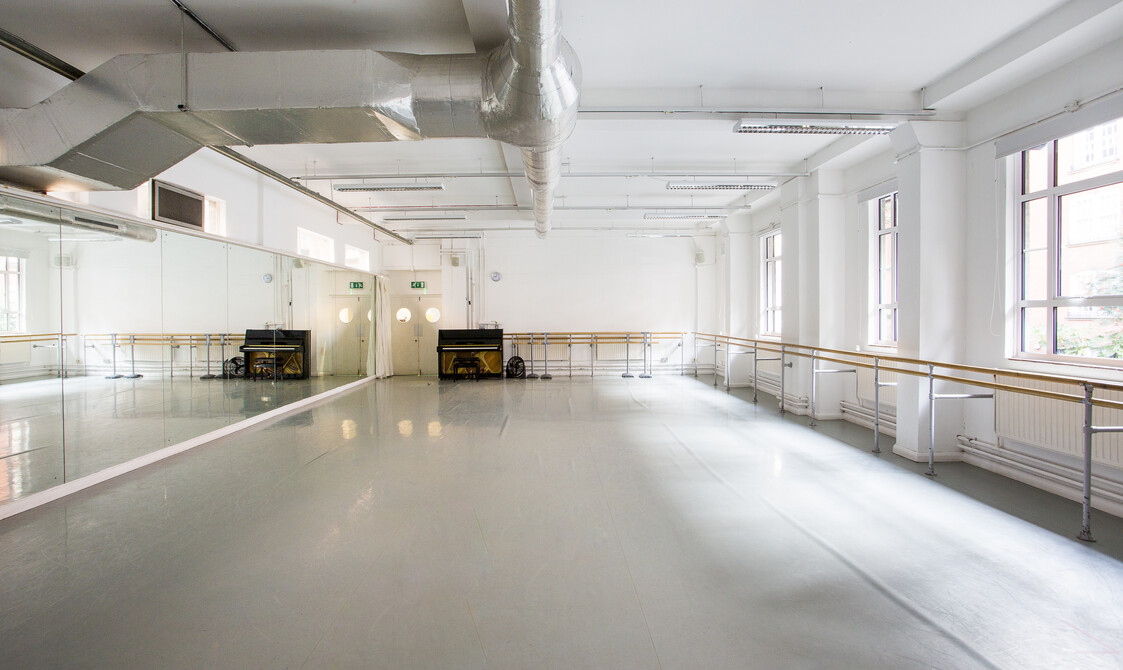 Image of an empty studio with windows on the right hand side with ballet barres across the wall, a piano on the far wall and mirrors on the left hand side