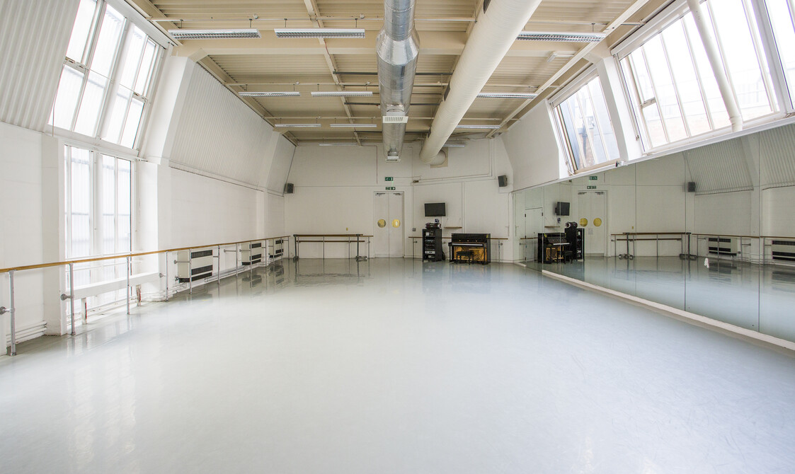 Image of an empty studio with windows and ballet barres on the left hand side, a piano by the far wall and mirrors on the right hand side