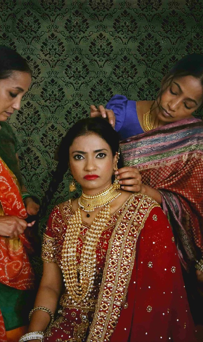 A woman wearing a sahri is looking down the camera and having her hair done by three other woman who are also wearing sahris. There is a decorative green wallpaper in the background.