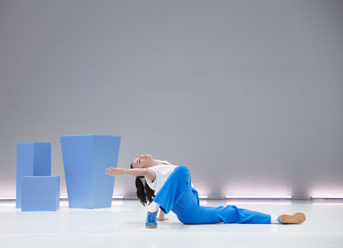 There is a white background and three blue cubes in the corner of the stage. A dancer is in the centre of the stage wearing a wearing top and blue trousers and trainers.