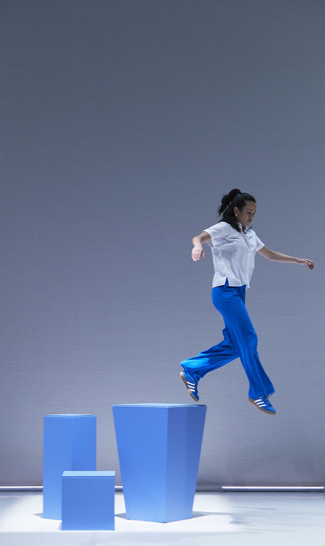 A dancer dressed in a white top and blue trousers jumps from a tall blue block.