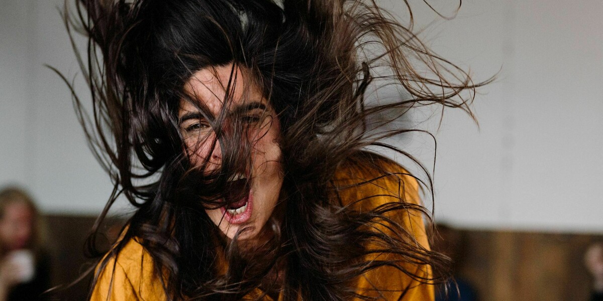 A woman wearing an orange shirt bangs her fist angrily on the table.