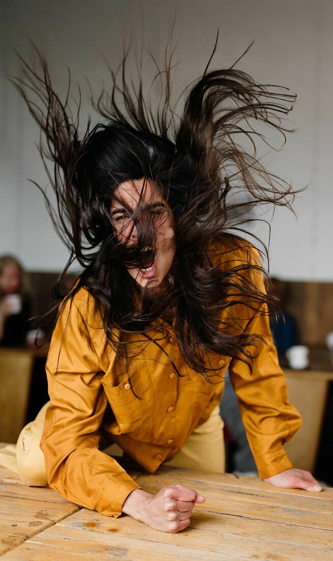A woman wearing an orange shirt bangs her fist angrily on the table.