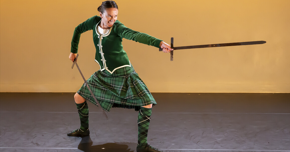 Person on stage wearing a green traditional Scottish outfit holding a sword in both hands and pointing one out.
