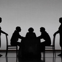 Black and white image of five bodies onstage. Three are sitting on a table facing each other. Two are standing behind the chairs that two of the bodies are sitting on.