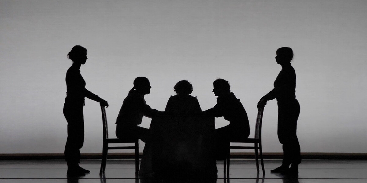Black and white image of five bodies onstage. Three are sitting on a table facing each other. Two are standing behind the chairs that two of the bodies are sitting on.