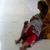 In the image the floor is white. A dancer dressed in yellow is kneeling on the floor. Another dancer wearing red Adidas stripe trousers and a grey top is lying in the other dancer's lap. They smile.