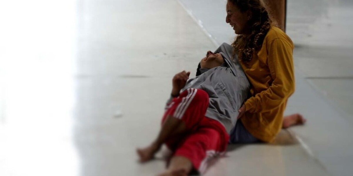 In the image the floor is white. A dancer dressed in yellow is kneeling on the floor. Another dancer wearing red Adidas stripe trousers and a grey top is lying in the other dancer's lap. They smile.