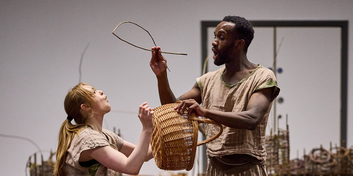 Two people stand facing each other holding a woven basket and a fish made from willow. They both look shocked and excited.