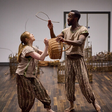 Two people stand facing each other holding a woven basket and a fish made from willow. They both look shocked and excited.