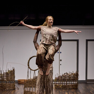 A man is facing away from us holding a woman on his shoulders. She is facing us holding a fish made from willow.