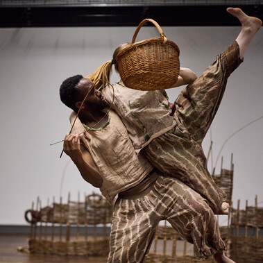 A man lifts a woman as she kicks her leg into the air. He is holding a fish made from willow and she is holding a woven basket.