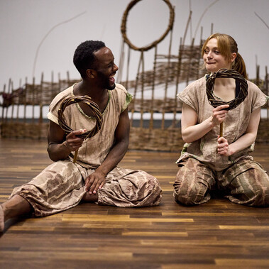 A man and woman sit on the floor holding willow rings and a stick. The man is laughing at the woman as she makes a funny face.