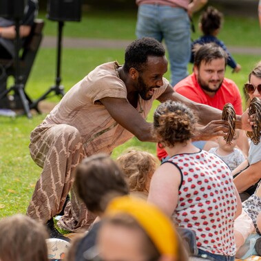 A man is kneeling next to an audience in a grassy park. People smile and laugh as he holds two willow rings towards a child.
