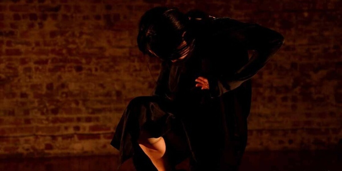 In the image there is a brick wall in the background. A dancer with black hair crouches over their body, lifting their right leg slightly so the foot is by the knee. The dancer is wearing all black.