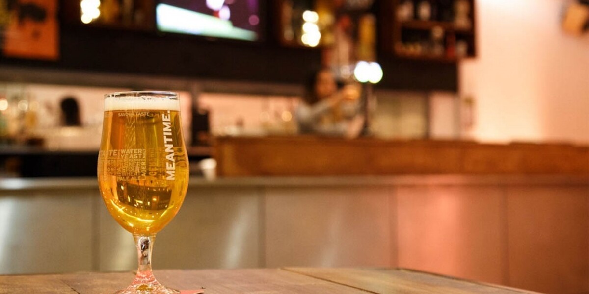 A photo of the theatre bar at The Place. We see a wooden table with a pint of meantime beer on it. The bar is in the background.