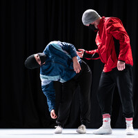 Two dancers in the image, one is leaning down to the right whilst the other looks on. One wears a blue jacket and a black hat with a mask, the other a red jacket, grey hat and a mask.