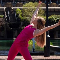 Image of Ellie dancing centrally in the camera. Behind Ellie is a water feature and a small tree. Ellie is wearing all pink clothes and has long blonde hair.