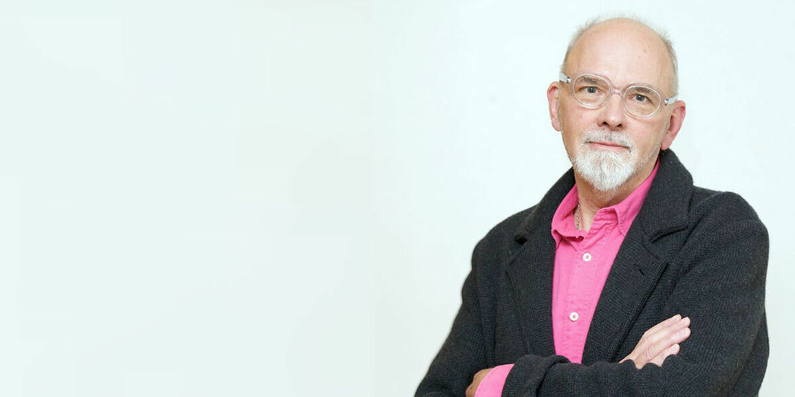 Image of Bob Lockyer. Bob looks into the camera with his arms folded. He is wearing a pink shirt with a black cardigan over the top.