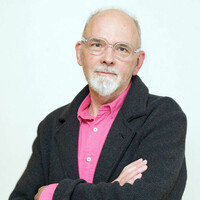 Image of Bob Lockyer. Bob looks into the camera with his arms folded. He is wearing a pink shirt with a black cardigan over the top.