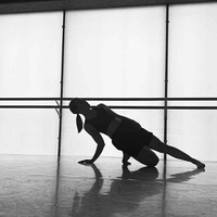 Black and white image of a dancer in the studio. A barre is in the background running through the image. There are also windows. The dancer kneels down on one leg with the other leg out. They also have their hand on the floor which, along with their knee, they are using to support themselves.