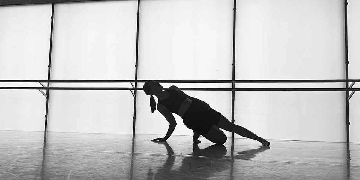 Black and white image of a dancer in the studio. A barre is in the background running through the image. There are also windows. The dancer kneels down on one leg with the other leg out. They also have their hand on the floor which, along with their knee, they are using to support themselves.