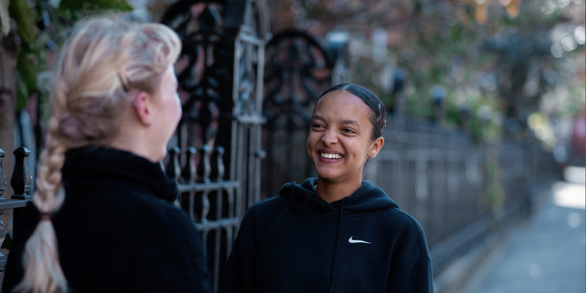 Two students stand outside LCDS, there is a blurred background that has a metal fence and trees in, one students is smiling at the other