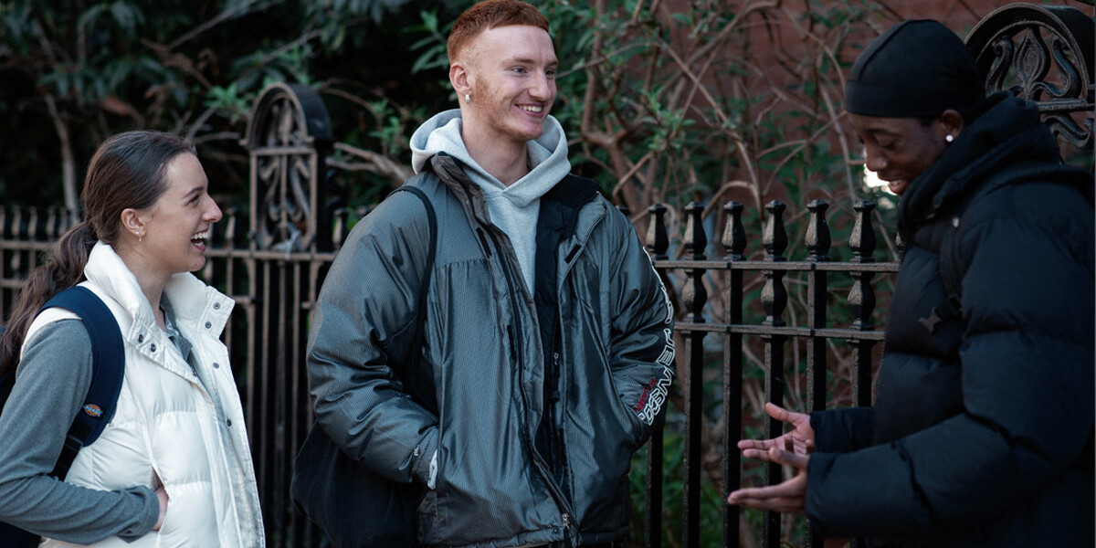 Three LCDS students stand outside The Place, there is a metal fence and trees behind them, one student is making the other two laugh