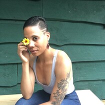 A woman with a grey strap top on covers her right eye with a yellow flower, she has tattoos on her left arm and short black hair