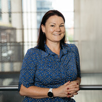 A white woman with dark hair up to her shoulders is facing us in front of a balcony, She is wearing a blue dress and is smiling.